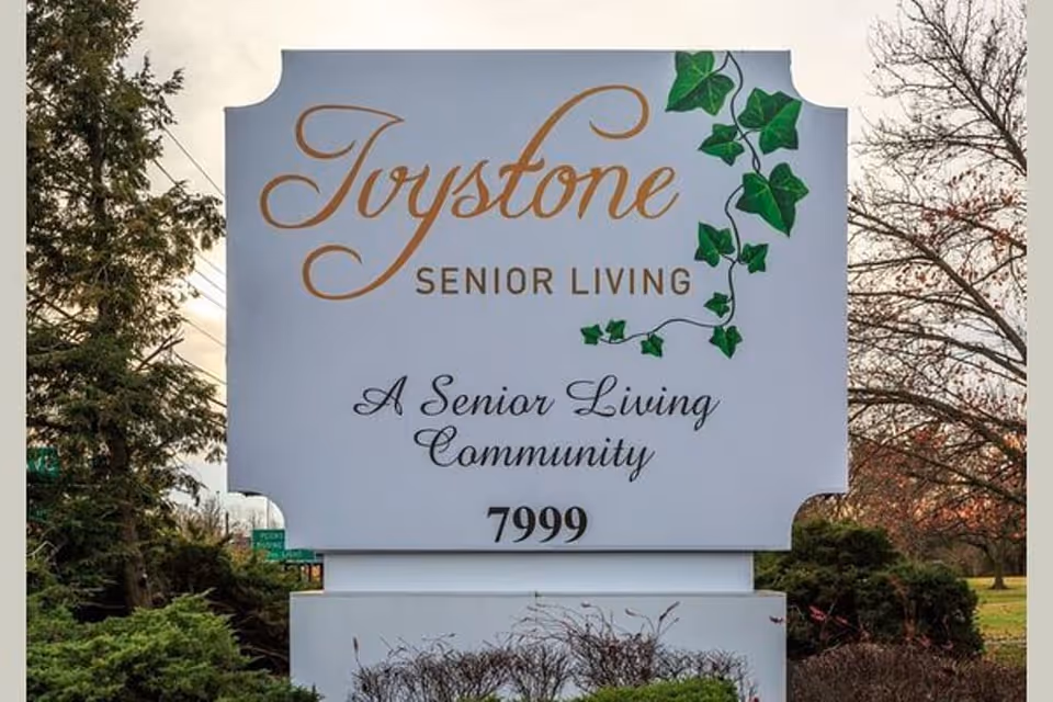 White outdoor sign reading 'Ivystone Senior Living - A Senior Living Community' with decorative ivy and the number 7999, set among trees and shrubs.