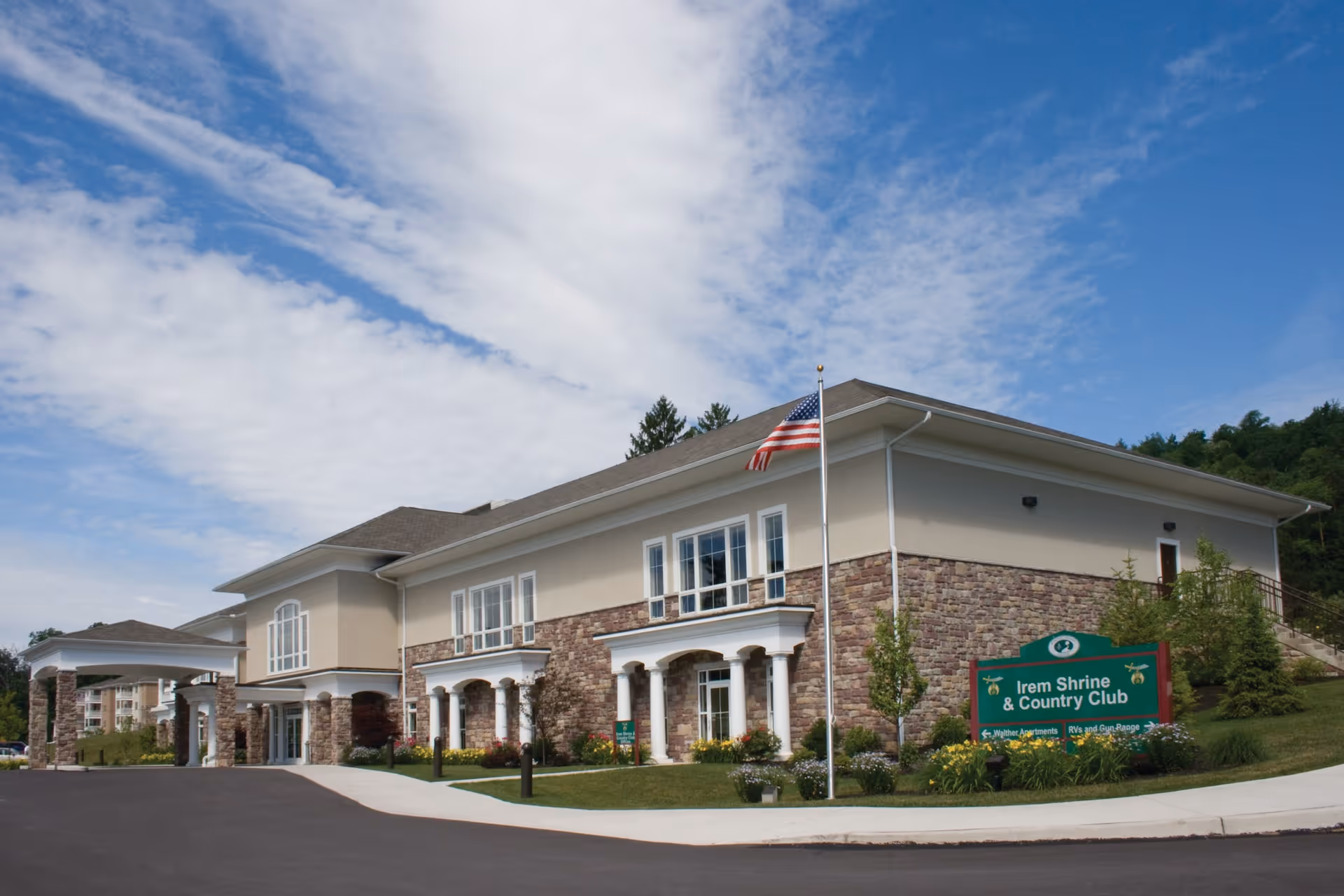 Exterior view of a large building with beige and stone facade, white columns, and an American flag on a flagpole in front. The building is identified as Irem Shrine & Country Club by a green sign near the entrance. The sky is partly cloudy with blue patches.