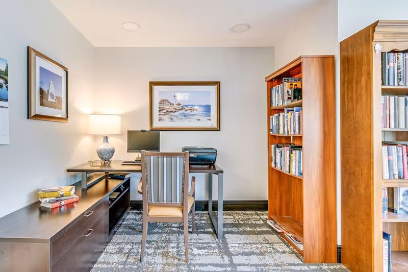 Small interior office/library space with a desk, chair, lamp, computer, printer, and bookshelves against the walls.