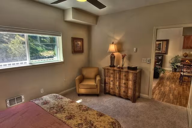 Sunlit bedroom with a bed and patterned comforter, an armchair, a wooden dresser with a lamp, and a window showing greenery outside.