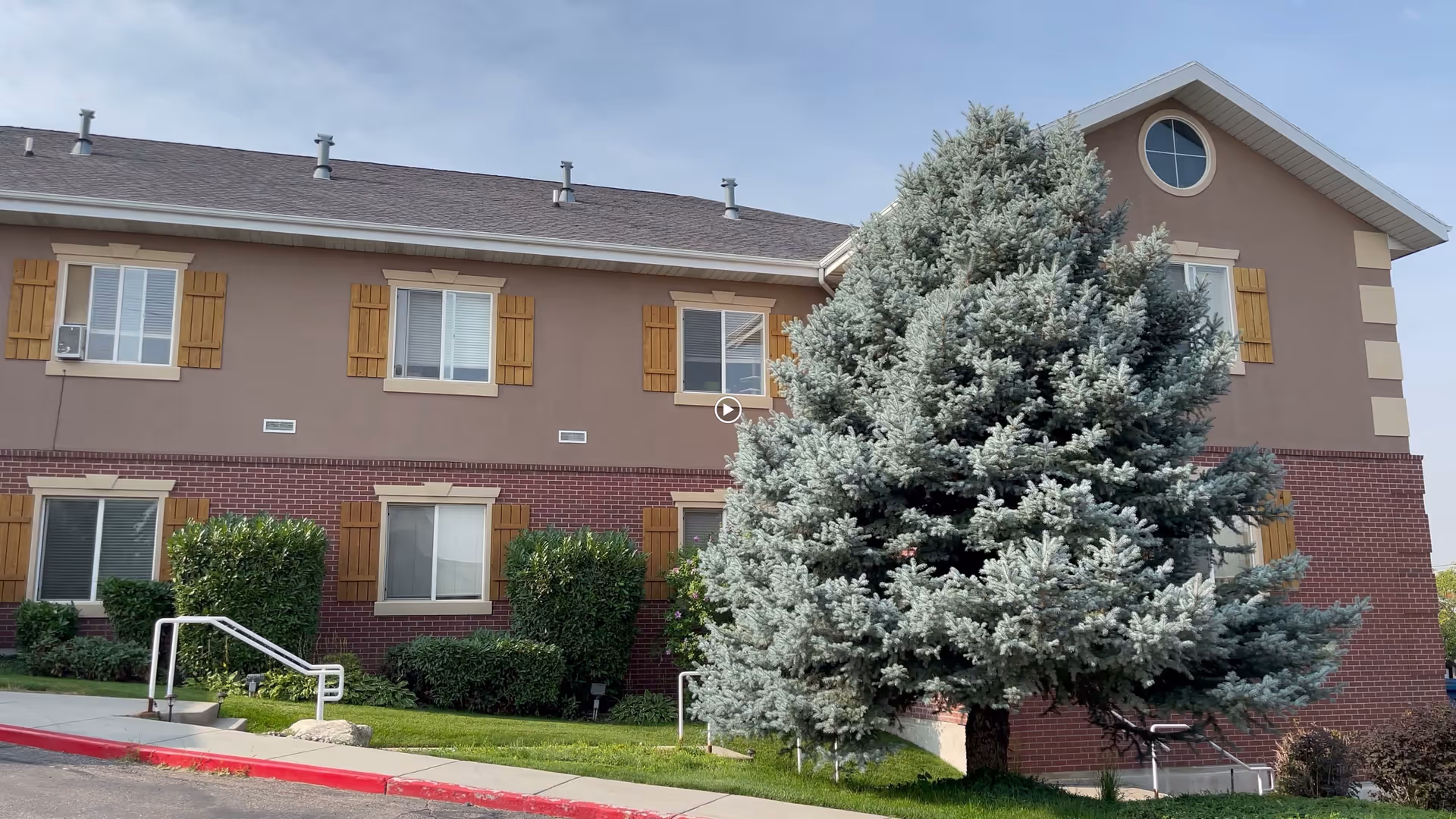 Exterior view of a two-story assisted living facility building with a combination of brick and beige siding. The building has several windows with wooden shutters and a large evergreen tree in front. There is a sidewalk and some green bushes along the building.