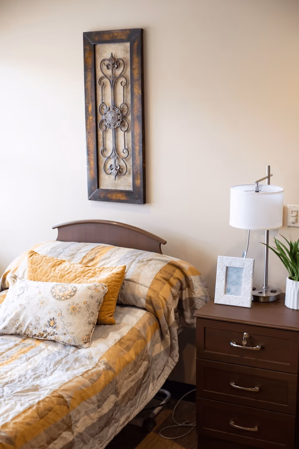 A neatly made bed with a yellow and beige striped quilt and two decorative pillows, one yellow and one floral patterned. Next to the bed is a wooden nightstand with three drawers, a white table lamp, a small potted plant, and a white photo frame. Above the bed hangs a decorative metal wall art piece with a rustic frame.