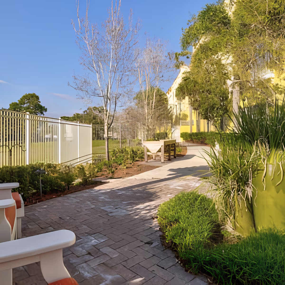 A paved outdoor walkway surrounded by greenery and trees, with a white bench on the left side and a wooden table further along the path. The sky is clear and blue.