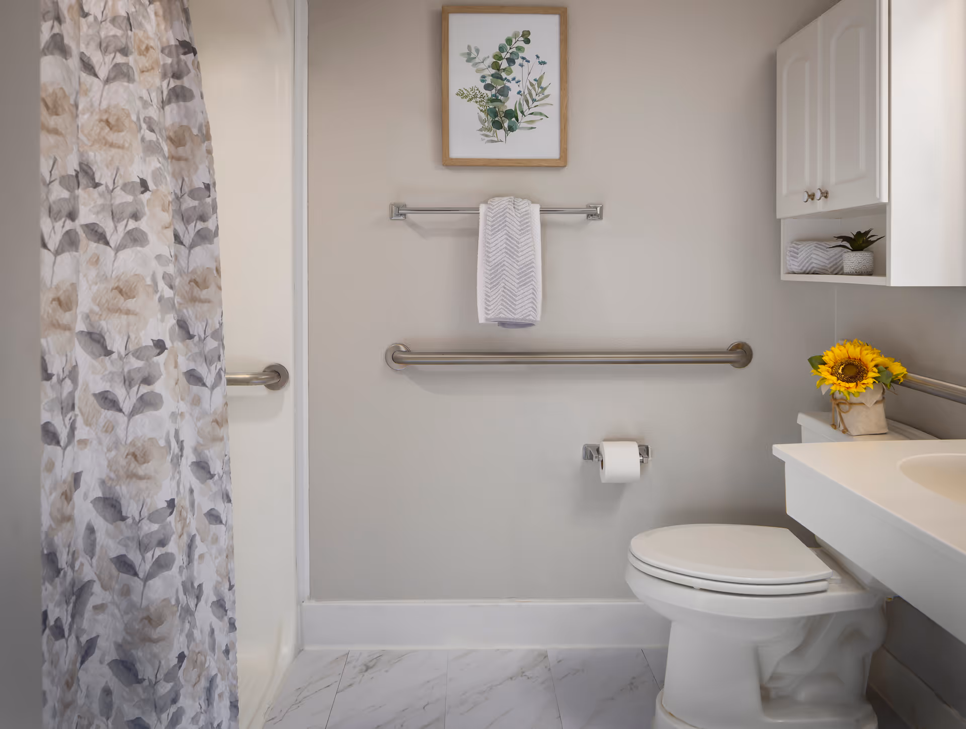 A clean and bright bathroom featuring a toilet, a white countertop with a sink, a cabinet with a small plant and a sunflower in a vase, a floral shower curtain, a towel hanging on a rack, and a framed botanical print on the wall.