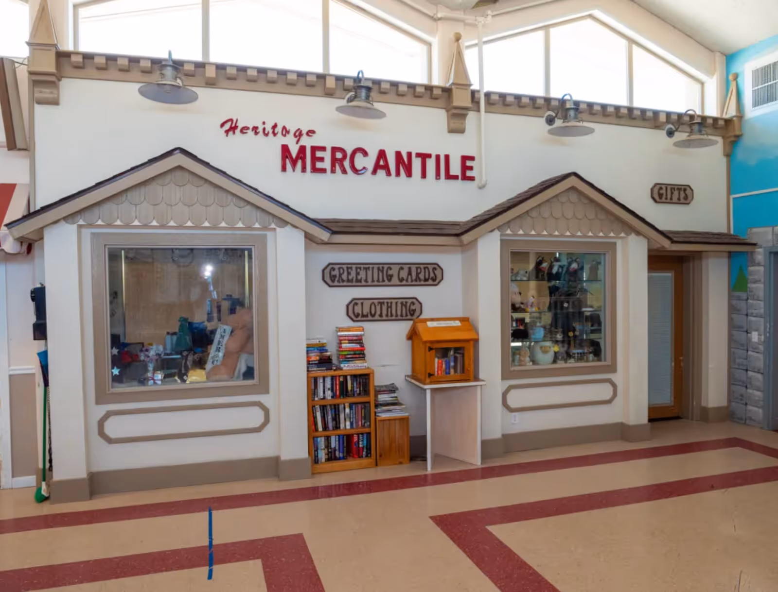 Interior view of a small indoor shop area named Heritage Mercantile with signs for greeting cards, clothing, and gifts. The shop has two display windows showing various items, a small bookshelf with books, and a donation or suggestion box on a stand. The floor has a beige and red pattern, and the ceiling has large windows letting in natural light.