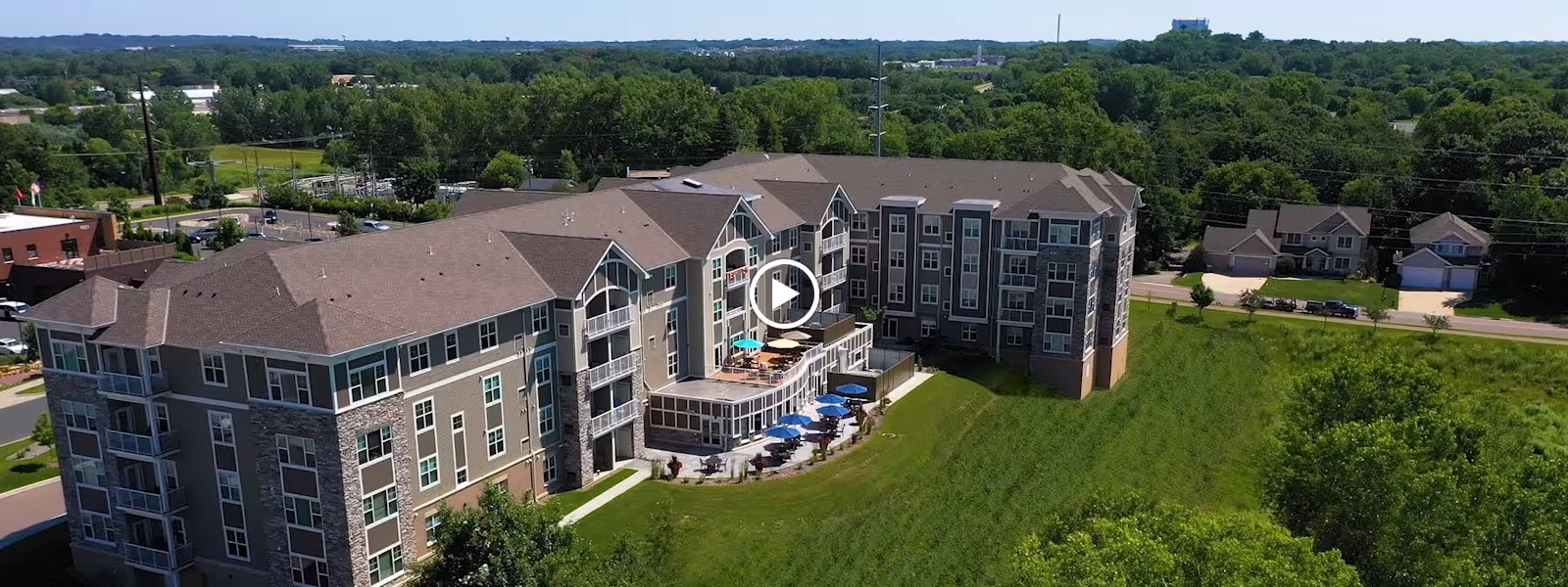Aerial view of a large senior living facility building with multiple floors, balconies, and outdoor patio areas with tables and umbrellas, surrounded by green lawns and trees, with a residential neighborhood and roads in the background.