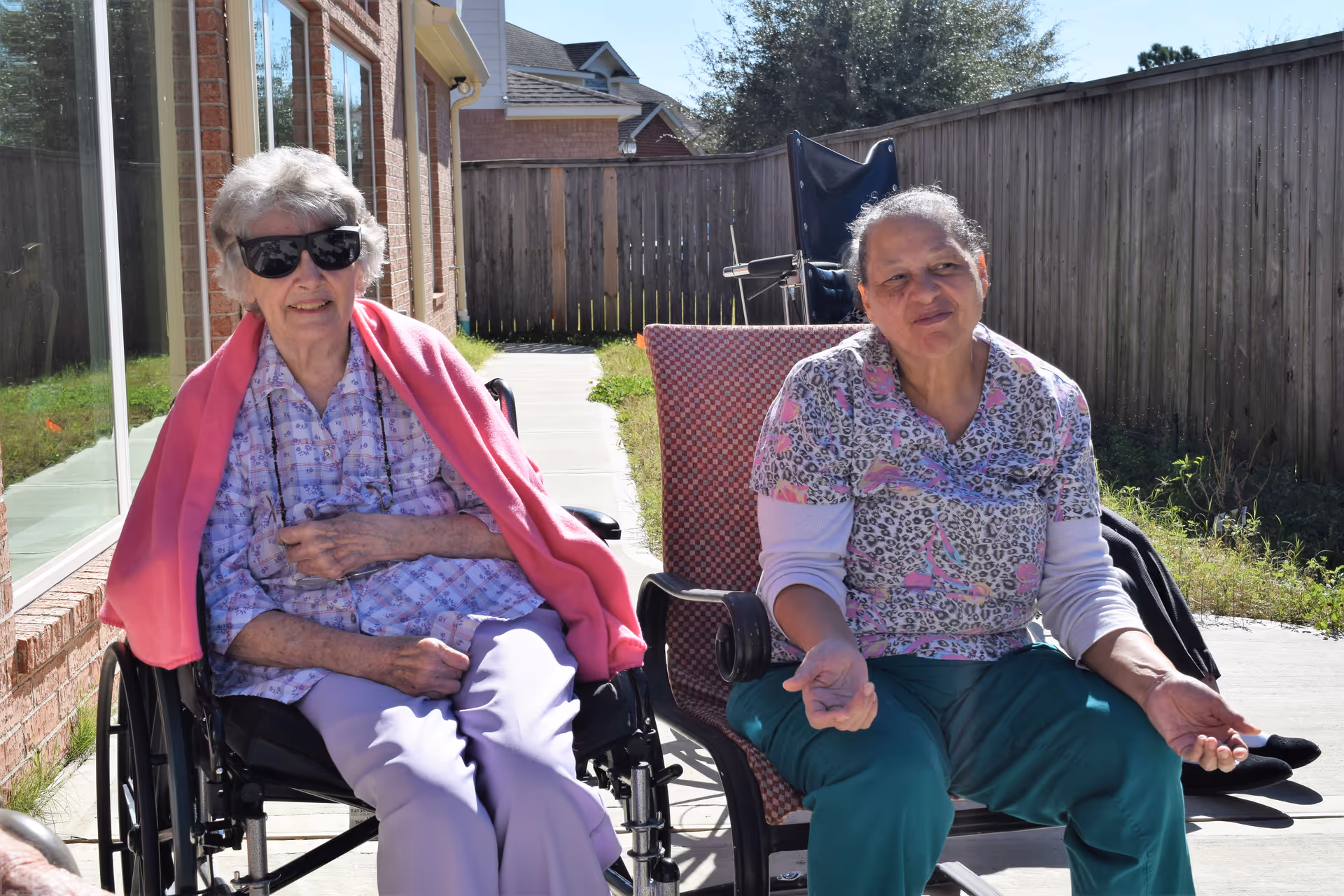 Two elderly women sitting outside on a sunny day. One woman is in a wheelchair wearing sunglasses, a pink shawl, and light purple pants. The other woman is sitting on a chair wearing a patterned shirt and green pants. They are next to a brick building with a wooden fence in the background.