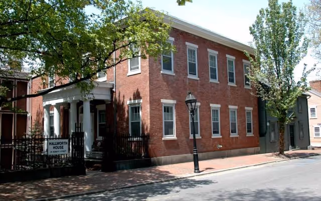 Exterior view of Hallworth House, a two-story brick building with white-trimmed windows and a white columned entrance. There are trees and a black iron fence in front, along with a streetlamp and a brick sidewalk.