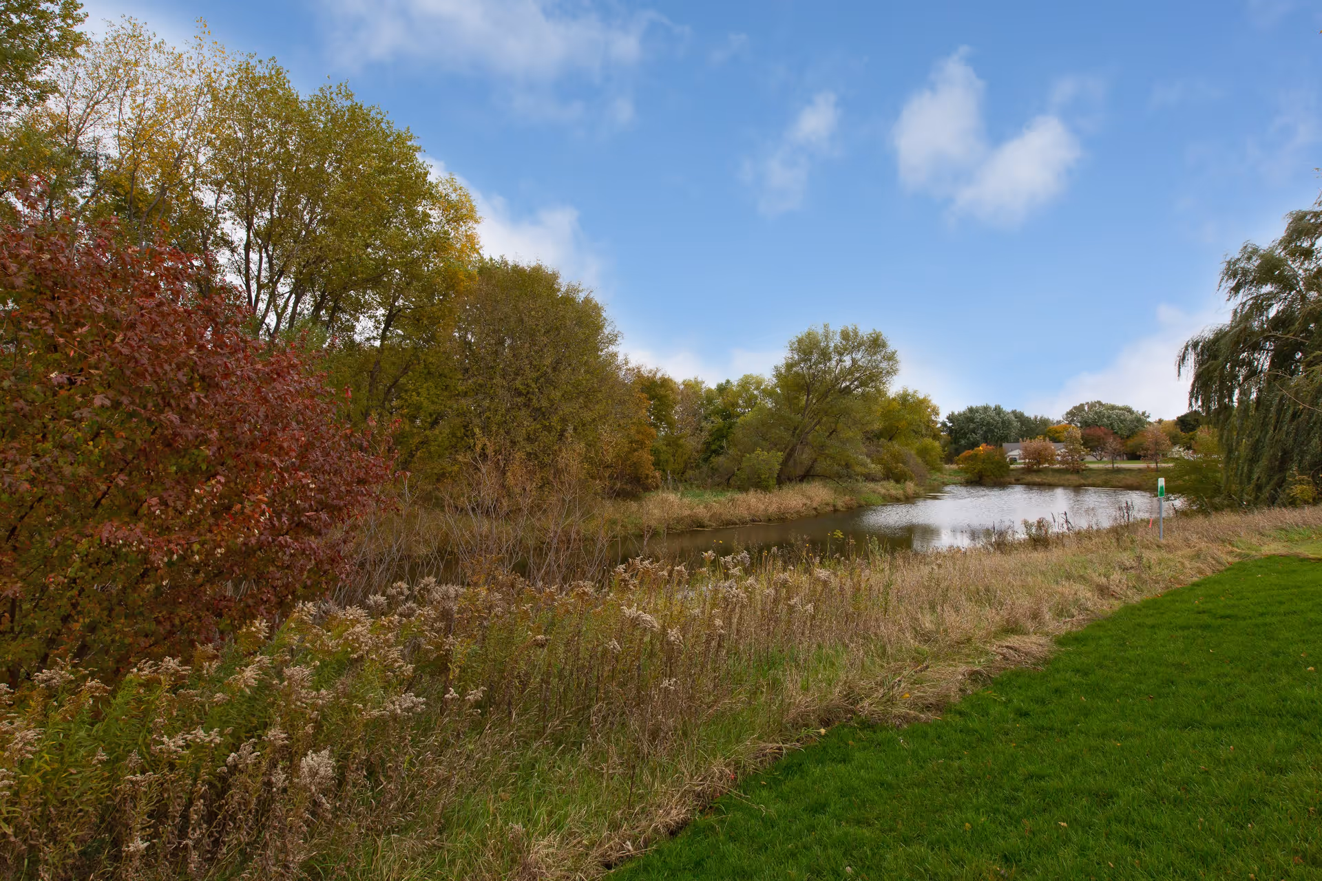 A scenic outdoor view featuring a small pond surrounded by trees with autumn foliage, tall grasses, and a well-maintained grassy area under a partly cloudy blue sky.