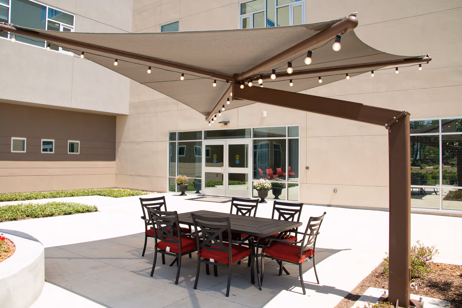 Outdoor courtyard patio with a dining table, red-cushioned chairs and a large shade canopy in front of a modern building entrance.