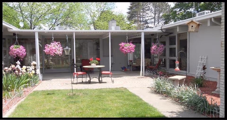 Outdoor patio area with a round table and four red cushioned chairs under a covered porch. Pink hanging flower baskets are suspended from the porch roof. The patio is surrounded by garden beds with flowers and greenery, and there is a grassy lawn in the foreground. The building exterior is light-colored with large windows.