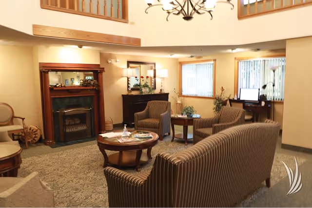 A warm common lounge with striped sofas and armchairs arranged around coffee tables in front of a fireplace and windows.