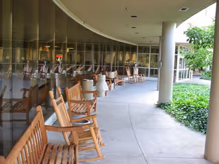 Curved outdoor walkway with wooden rocking chairs lined up along a glass wall on one side and greenery on the other side, supported by large white columns.