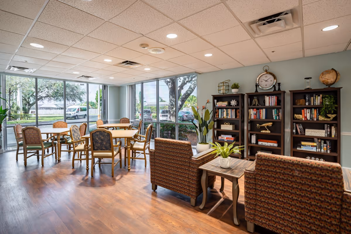 A bright and spacious common area with large windows showing an outdoor view. The room features several wooden tables with chairs arranged around them, two patterned armchairs facing a small wooden table with a potted plant, and three dark wooden bookshelves filled with books and decorative items including a clock, globe, and plants. The floor is wooden, and the ceiling has recessed lighting.