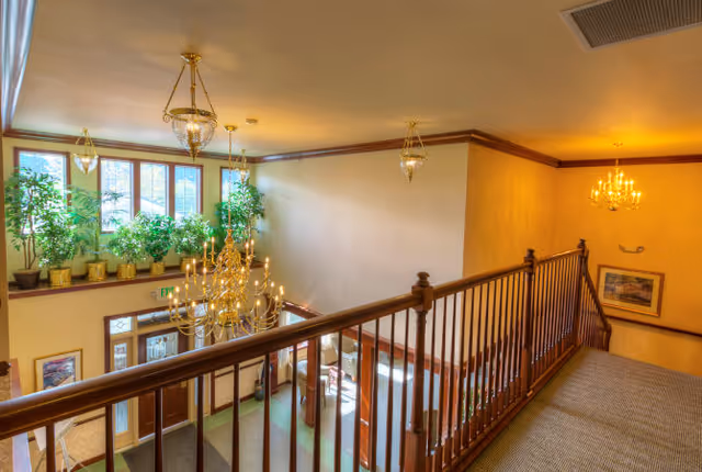 Second-floor interior balcony overlooking a lobby with wooden railings, chandeliers, and potted plants by tall windows.