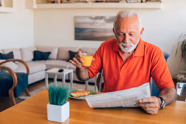 An elderly man with white hair and beard wearing a red polo shirt is sitting at a wooden table in a living room. He is holding a glass of orange juice in one hand and reading a newspaper with the other. On the table, there is a small potted plant and a plate with a croissant. In the background, there is a beige sofa with cushions and a coffee table.