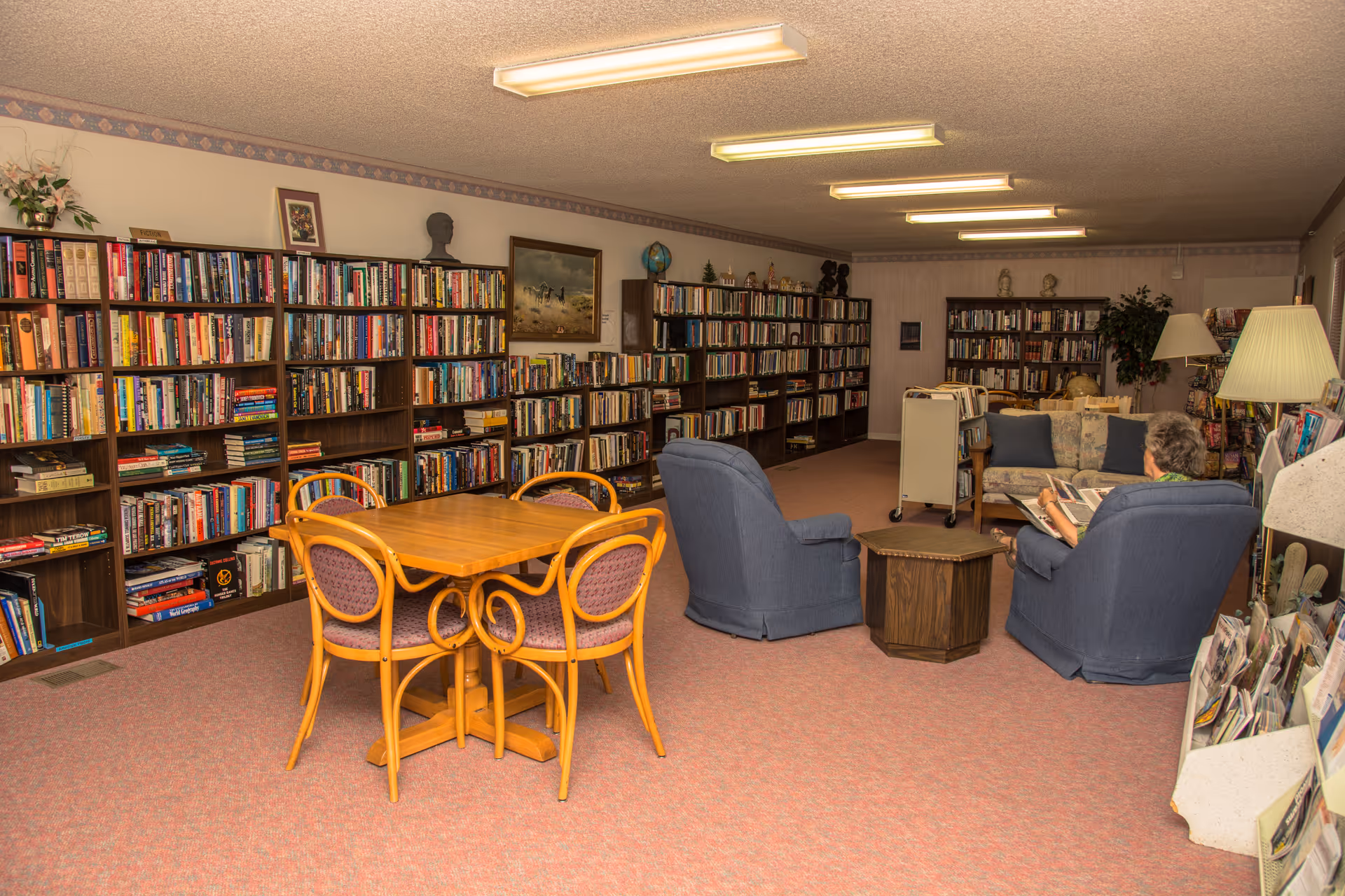 A cozy library or reading room with multiple bookshelves filled with books along the walls. There is a wooden table with four chairs in the foreground and a seating area with two blue armchairs and a sofa where a person is sitting and reading. The room is warmly lit with ceiling lights and has carpeted flooring.