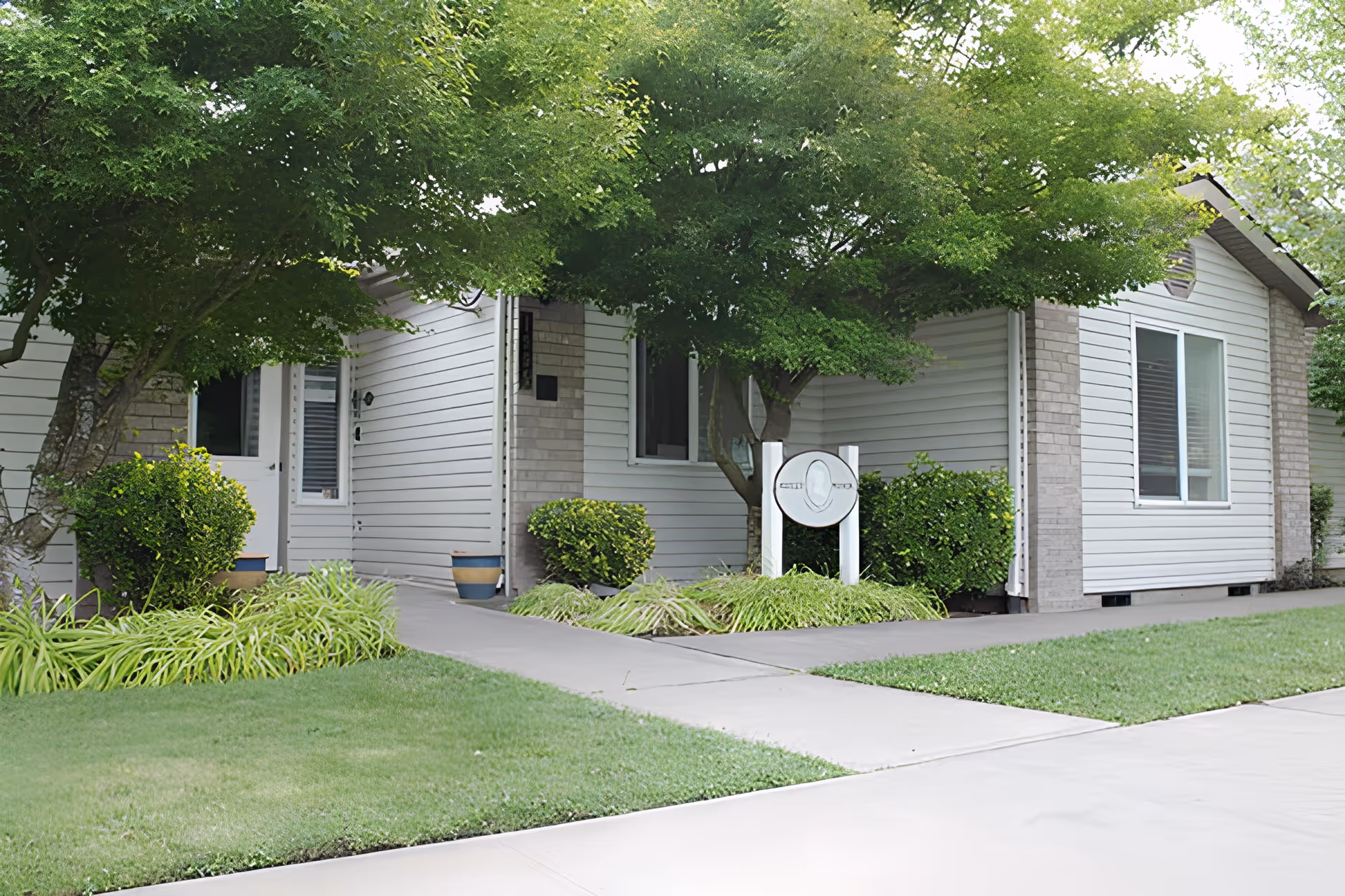 Exterior view of a single-story building with white siding and brick accents, surrounded by green trees, bushes, and a well-maintained lawn. A concrete walkway leads to a door, and there is a white signpost with a circular emblem near the entrance.