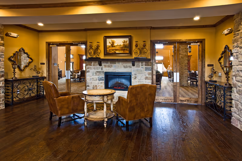 Interior lounge with two leather armchairs and a small round table facing a stone fireplace flanked by glass double doors.