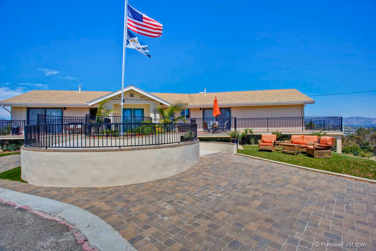 Front exterior of a single-story senior living building with a flagpole, patio seating and a paved driveway.