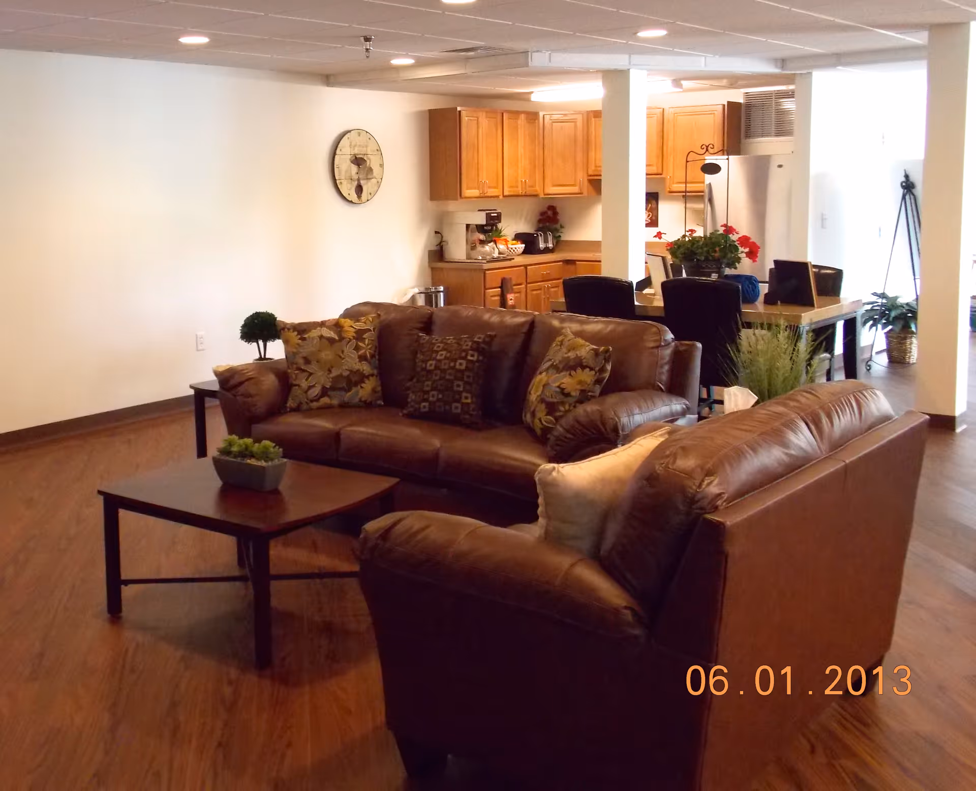 Interior view of a senior living facility common area with two brown leather sofas arranged around a wooden coffee table with a small plant. In the background, there is a kitchen area with wooden cabinets, a refrigerator, and a dining table with chairs. The floor is wooden, and the walls are light-colored with a clock hanging on one wall.