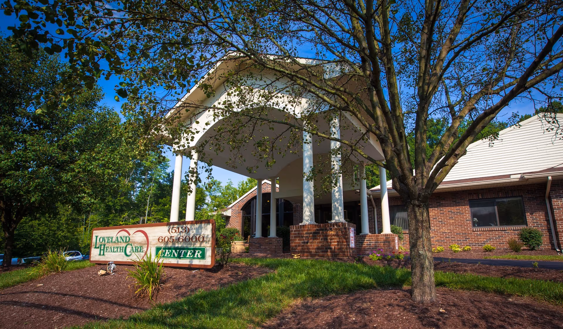 Exterior view of Loveland Health Care Center building with a covered entrance supported by white columns, surrounded by trees and landscaping under a clear blue sky.