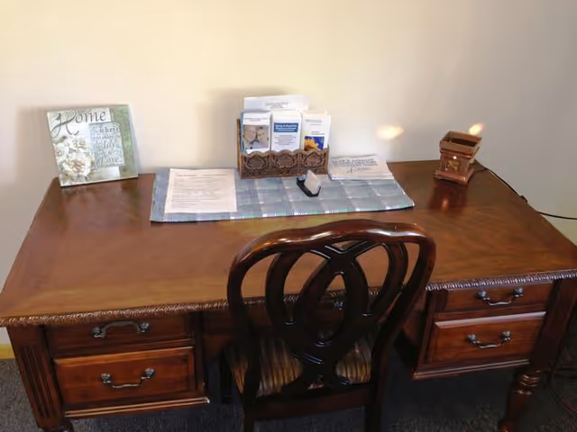 Wooden desk with a carved chair holding pamphlets, papers, and a small lamp against a plain wall.
