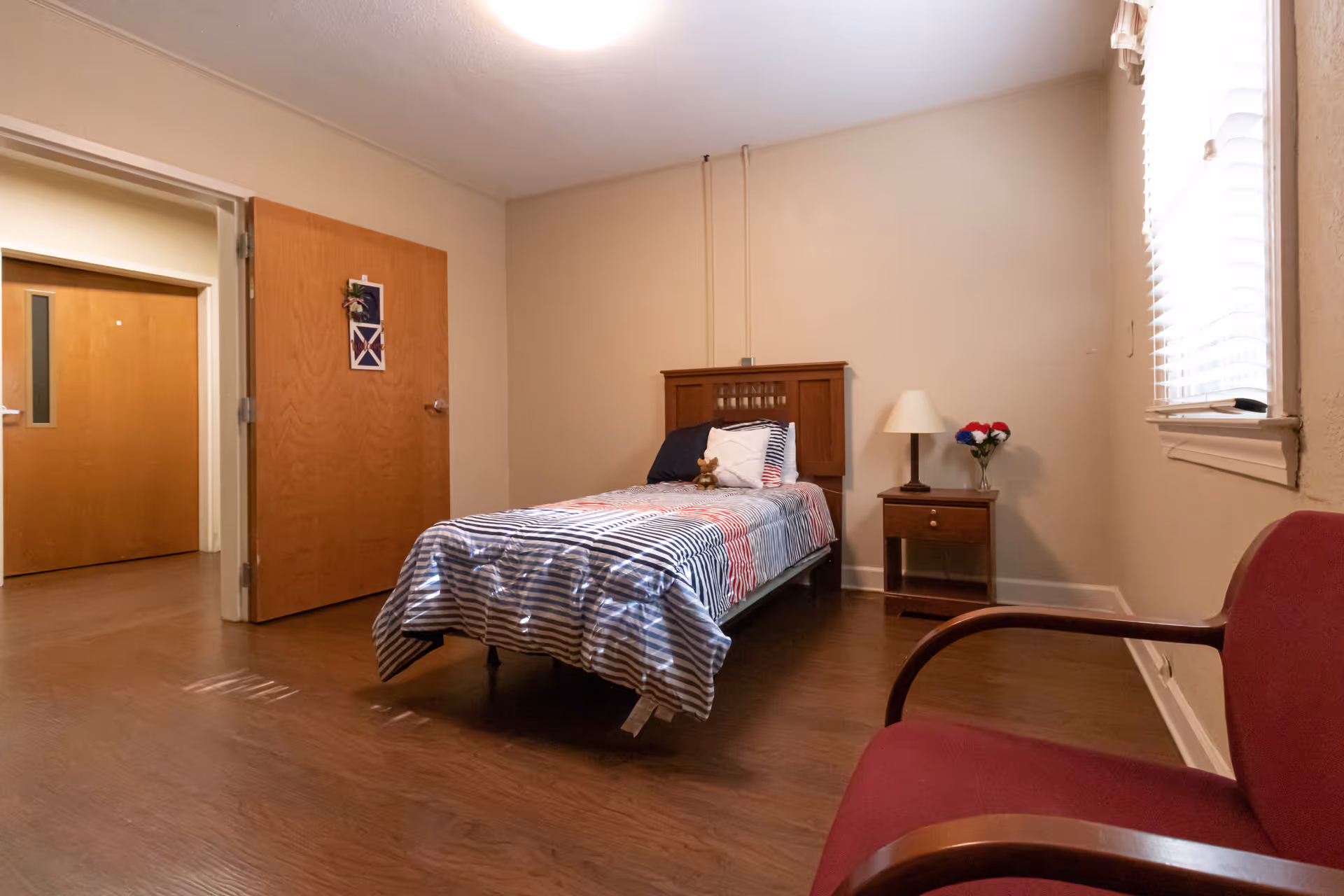 A simple bedroom with a single bed covered in a striped bedspread, a wooden headboard, a nightstand with a lamp and a vase of flowers, a red armchair, and a window with blinds letting in natural light. The room has wooden flooring and beige walls.