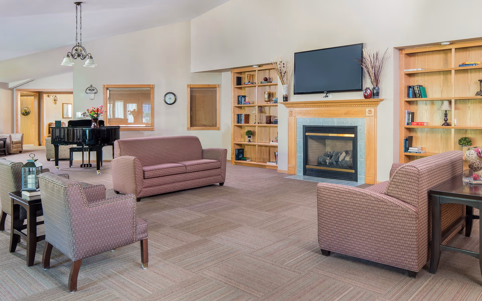 A spacious living room area with beige walls and carpeted floor, featuring two patterned sofas and two armchairs arranged around a fireplace with a mounted flat-screen TV above it. Built-in wooden bookshelves flank the fireplace, holding books and decorative items. A black grand piano is visible in the background near a window, and a hanging light fixture is suspended from the ceiling.