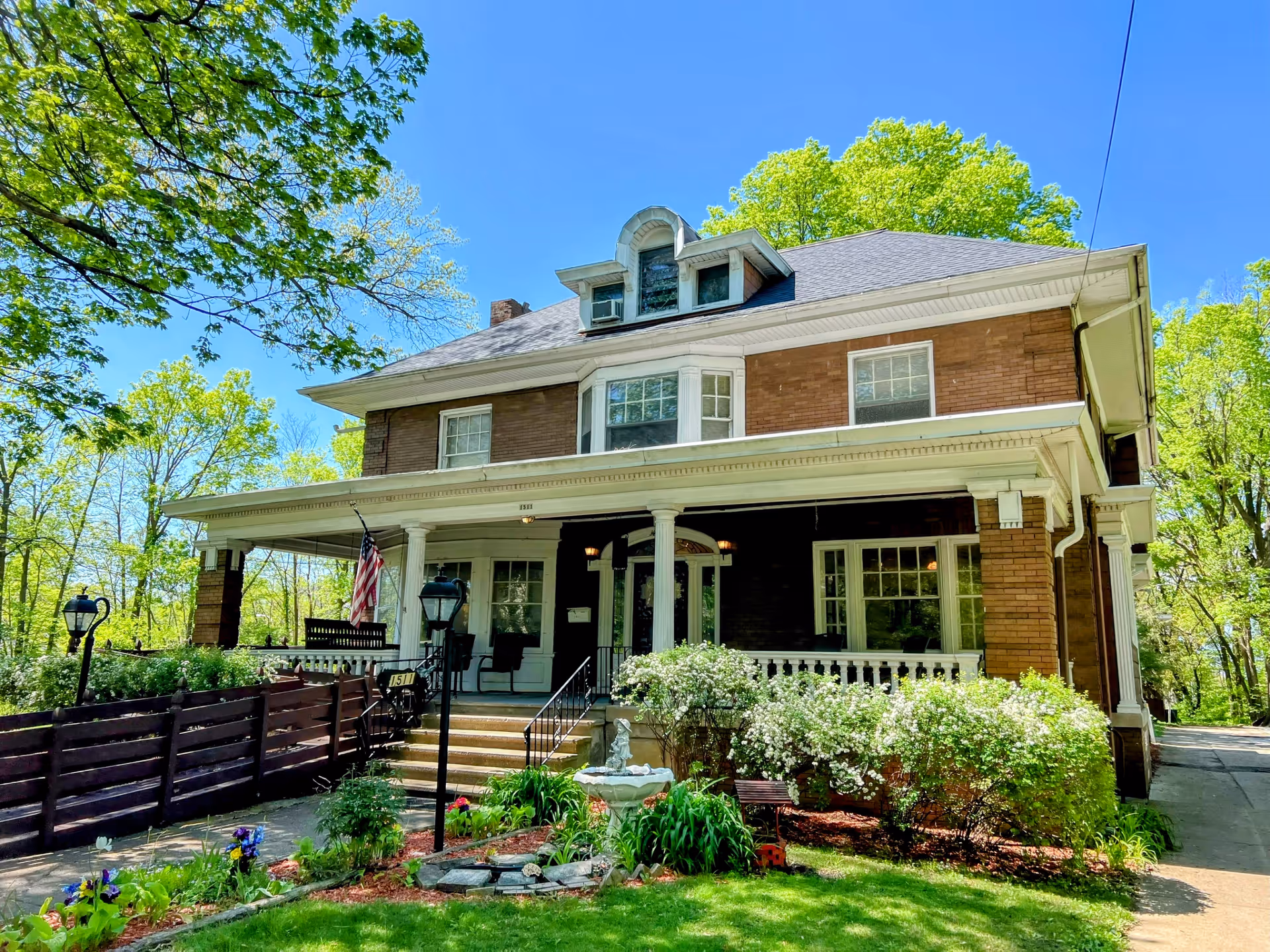 Front view of a large two-story brick house with a wraparound porch, landscaped yard, and an American flag.