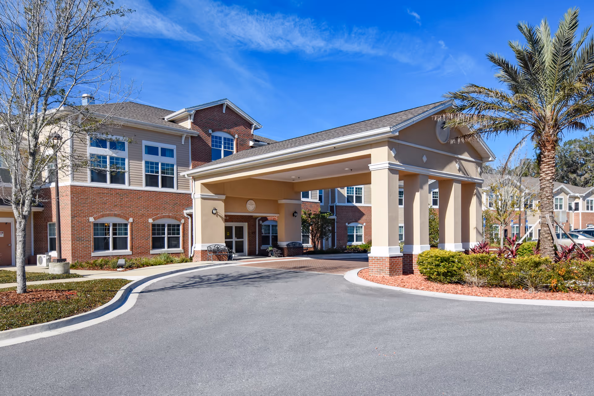 Exterior view of Anthem Lakes senior living facility showing a covered entrance with columns, a circular driveway, landscaped greenery including a palm tree, and a multi-story brick and beige building under a clear blue sky.