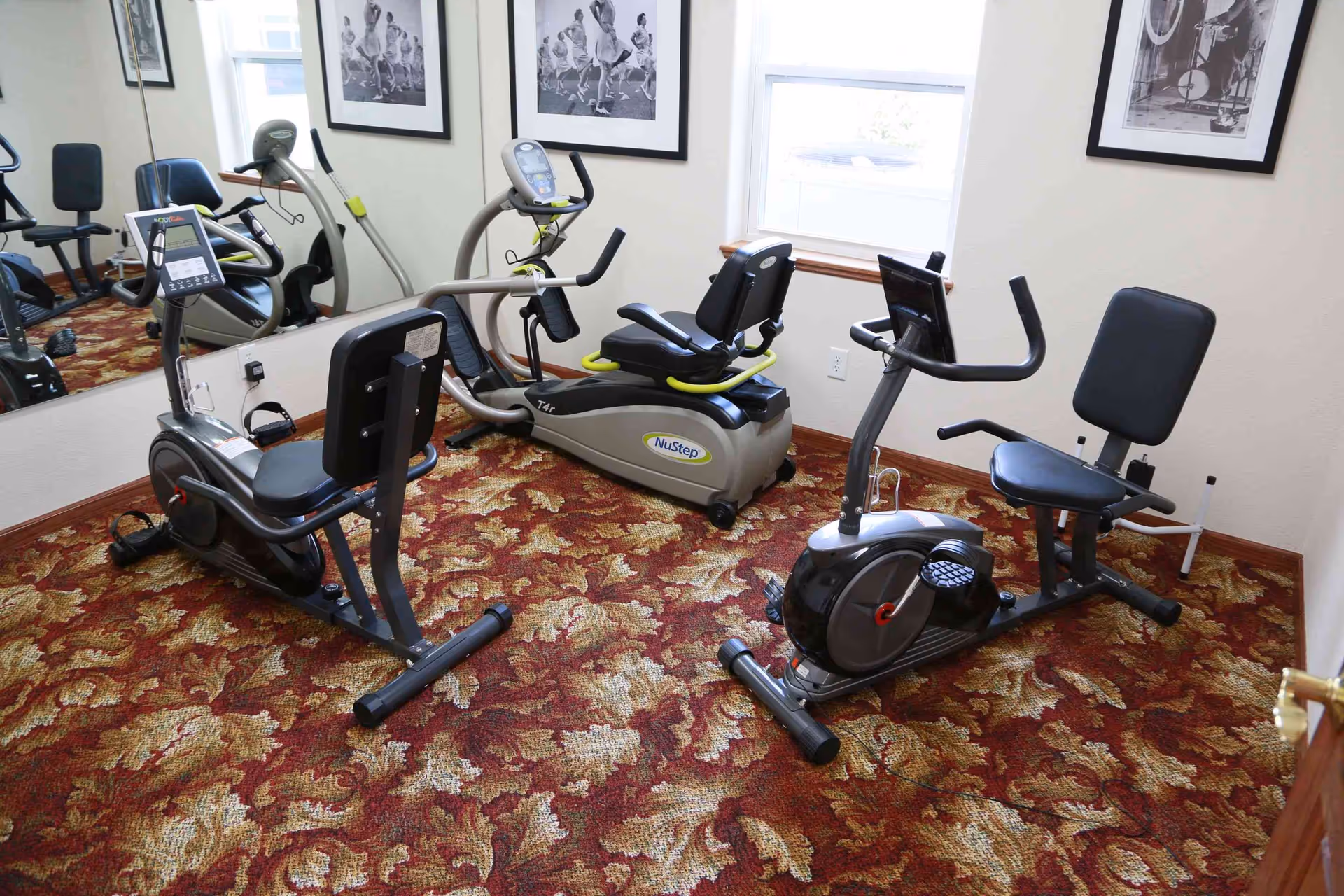 Small fitness room with three recumbent exercise bikes on patterned carpet, a mirrored wall and framed photos.