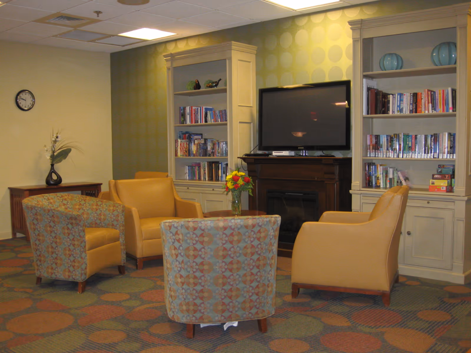 A cozy living room area with four armchairs arranged around a small round table with a vase of flowers. Behind the chairs are two built-in bookshelves filled with books and decorative items, and a flat-screen TV mounted above a fireplace. The room has patterned carpet and a wall clock on the left wall.