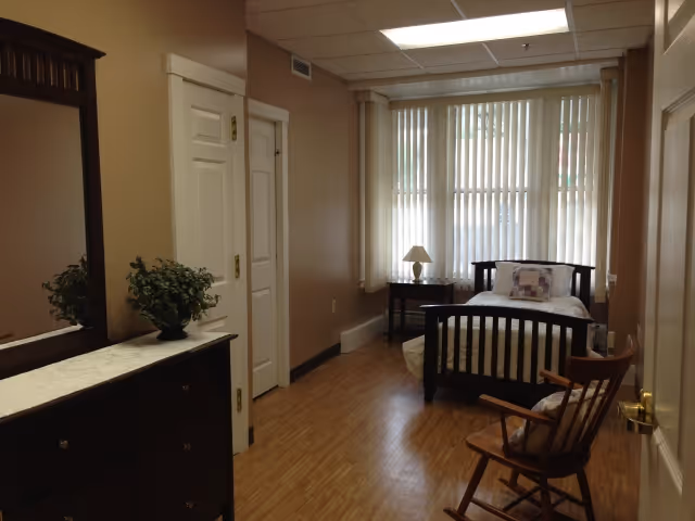 A senior living facility bedroom with a single wooden bed, a small bedside table with a lamp, a wooden rocking chair with a cushion, and a dresser with a mirror and a potted plant. The room has large windows with vertical blinds and wood flooring.