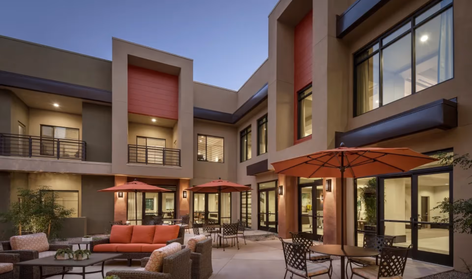Outdoor courtyard area of a senior living facility at dusk with comfortable seating including cushioned chairs and sofas, round tables with umbrellas, and modern two-story building with large windows surrounding the space.