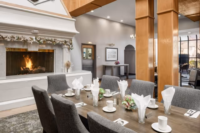 A dining area in a senior living community featuring a wooden table set for six with white napkins folded in glasses, cups, saucers, and silverware. There is a lit fireplace with a decorative garland above it on the left side, and wooden pillars and large windows are visible in the background.