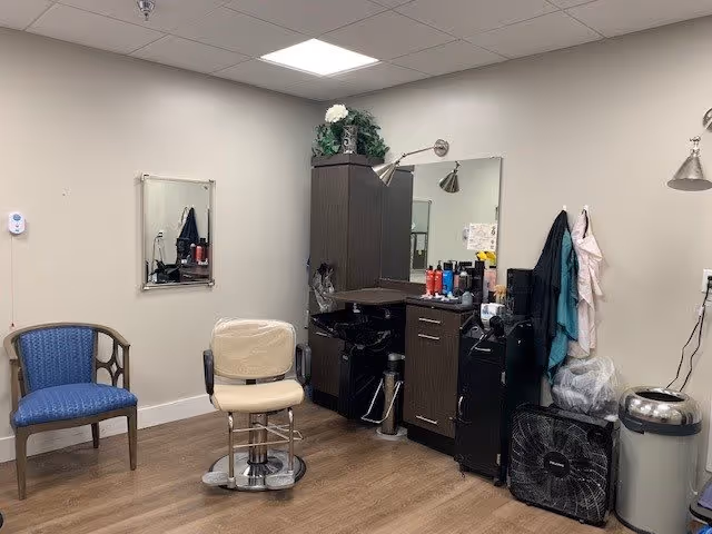 Interior of a hair salon area within a senior living facility, featuring a salon chair, a large mirror, a cabinet with hair care products, a blue guest chair, and various salon tools and equipment against beige walls and wood flooring.
