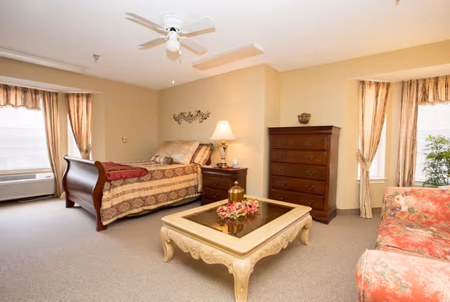 A cozy senior living bedroom with a wooden bed dressed in patterned bedding, a nightstand with a lamp, a tall wooden dresser, a floral upholstered sofa, and a decorative coffee table with flowers. The room has beige walls, carpeted floor, two windows with curtains, and a ceiling fan.
