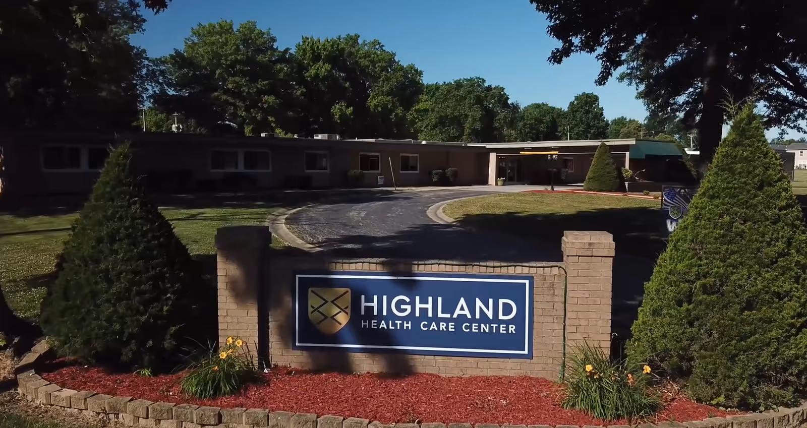 Front exterior view of Highland Health Care Center building with a curved driveway, surrounded by green trees and landscaped bushes with red mulch.