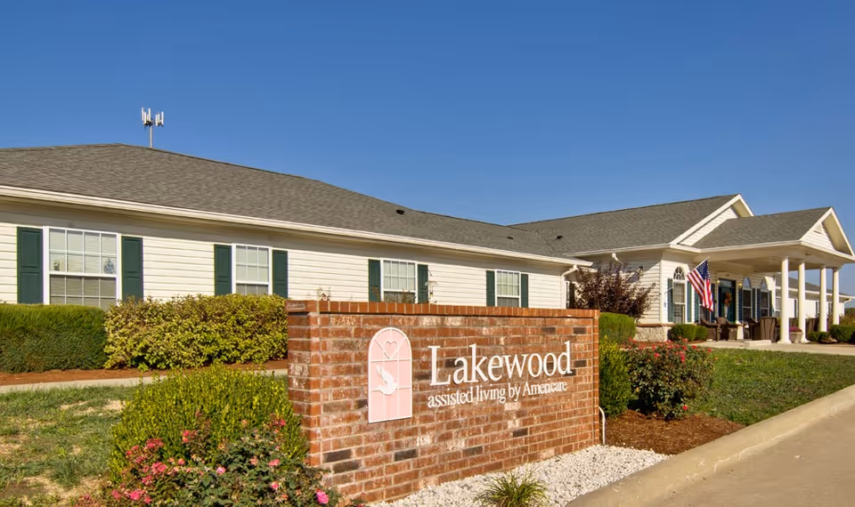 Brick sign reading 'Lakewood assisted living by Americare' sits in landscaped grounds in front of a single-story senior living building with a columned entrance and American flags.