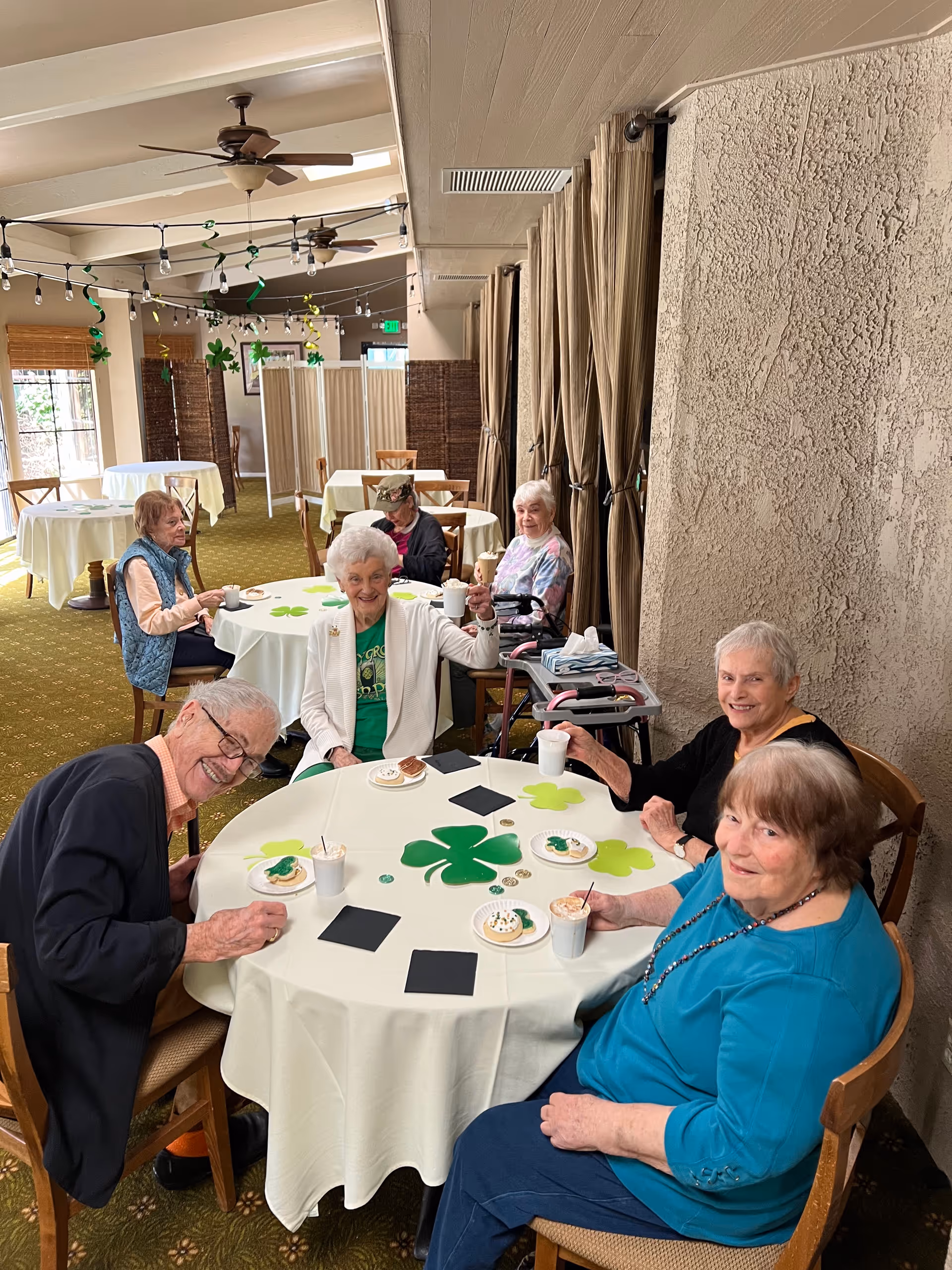 A group of elderly people sitting around a round table in a decorated dining area, enjoying drinks and cookies. The table is covered with a white tablecloth and decorated with green shamrocks and gold coins, suggesting a St. Patrick's Day celebration. The room has ceiling fans, string lights, and several other tables with chairs.