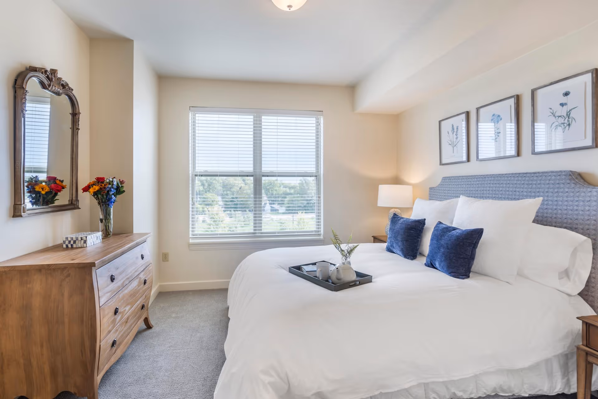 A bright and cozy bedroom with a large bed dressed in white linens and blue accent pillows. Above the bed are three framed botanical prints. To the left, there is a wooden dresser with a decorative mirror above it and a vase of colorful flowers. A window with blinds lets in natural light, illuminating the room.