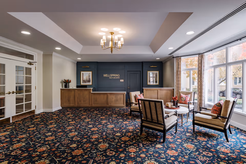 A spacious and well-lit reception area with a patterned carpet, a chandelier, and a seating arrangement of four cushioned chairs around a round table. The reception desk is wooden with a dark blue backdrop that has the text 'WELLSPRING HEALTHCARE CENTER'. Large windows on the right side let in natural light and show autumn trees outside.