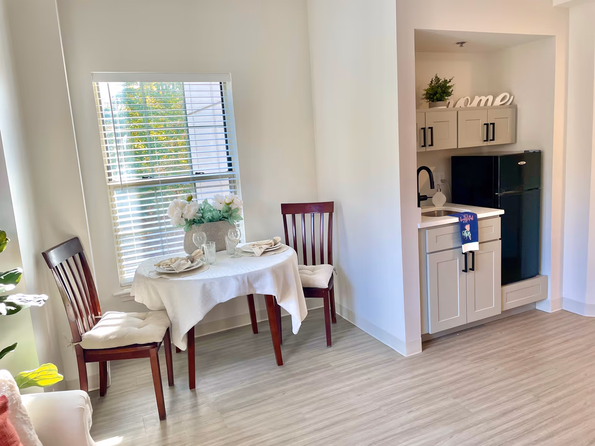 A small dining area with a round table covered with a white tablecloth, set with two place settings including plates, napkins, and glasses. Two wooden chairs with cushions are placed around the table. Next to the dining area is a compact kitchenette with light gray cabinets, a small sink, a black mini refrigerator, and a decorative plant on top of the cabinets. A window with white blinds allows natural light into the room.