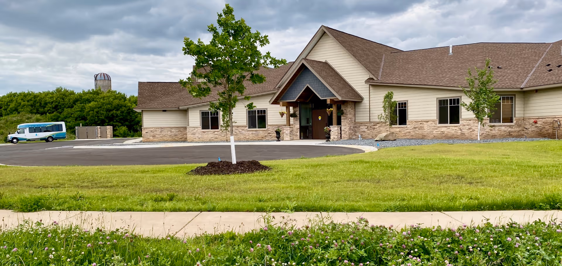 Single-story senior living building with a covered main entrance, circular driveway, green lawn and a parked shuttle van under a cloudy sky.