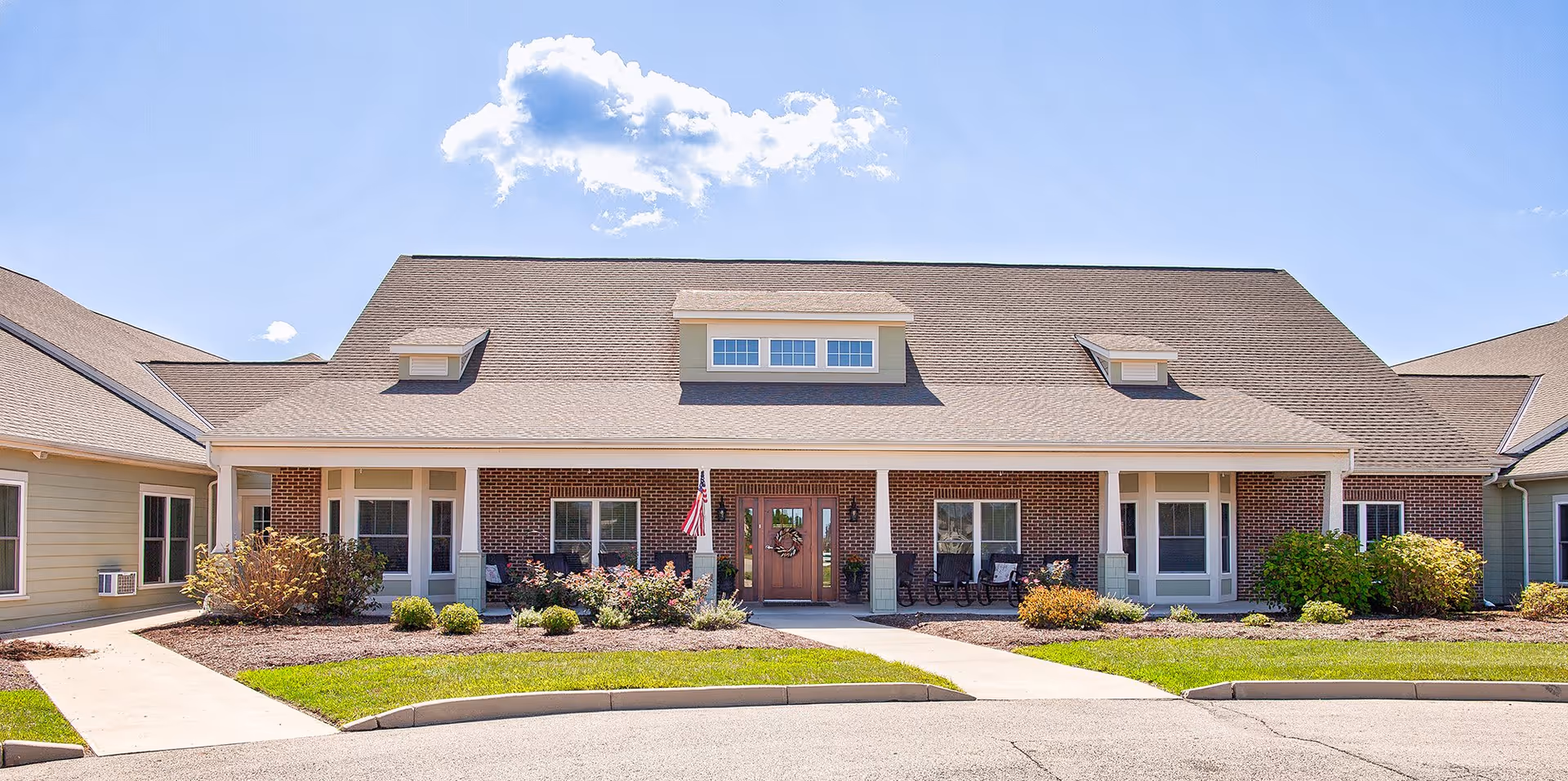 Front exterior view of a single-story senior living facility building with a brick facade, multiple windows, a central entrance door adorned with a wreath, and a small porch area with rocking chairs. The building is surrounded by landscaped bushes and a neatly maintained lawn under a clear blue sky with a few clouds.