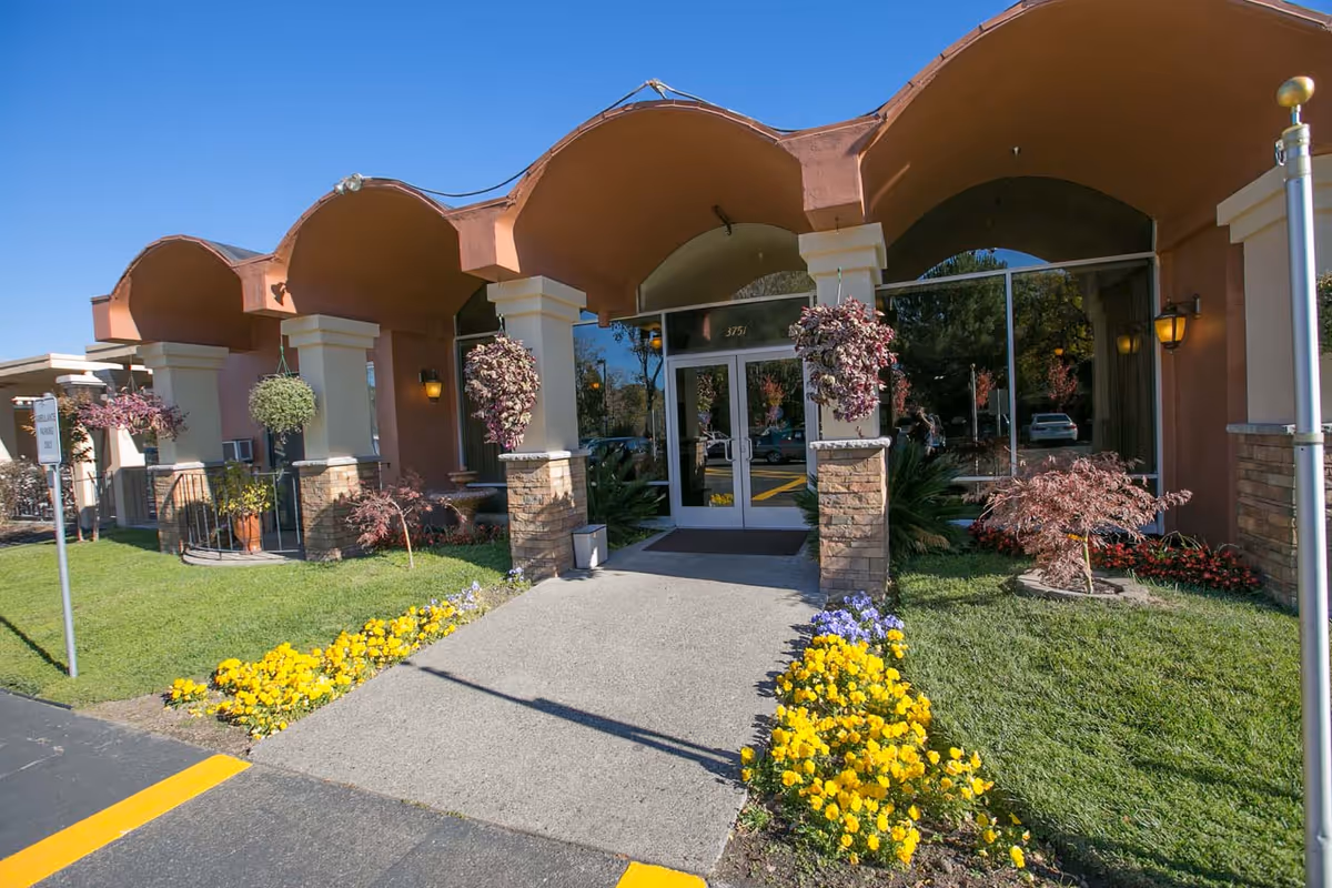 Front entrance of a stucco building with an arched canopy, glass double doors, hanging plants, and flowerbeds.