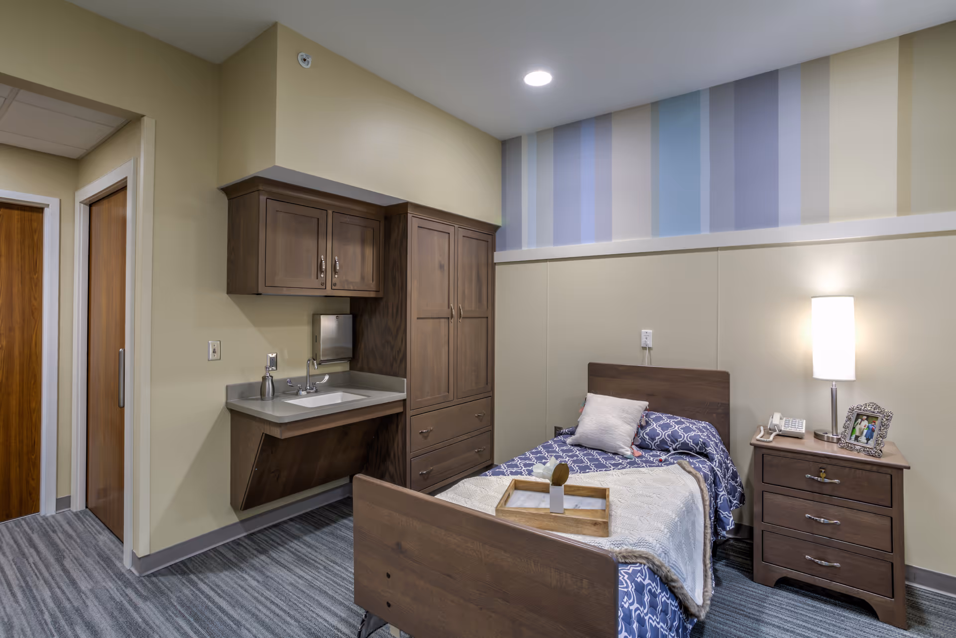 A single bedroom in a nursing and rehabilitation facility featuring a wooden bed with blue and white bedding, a wooden nightstand with a lamp, telephone, and framed photo, a built-in sink with cabinets above and below, and a door leading to another room. The walls are painted beige with a striped accent near the ceiling, and the floor is covered with gray carpet.