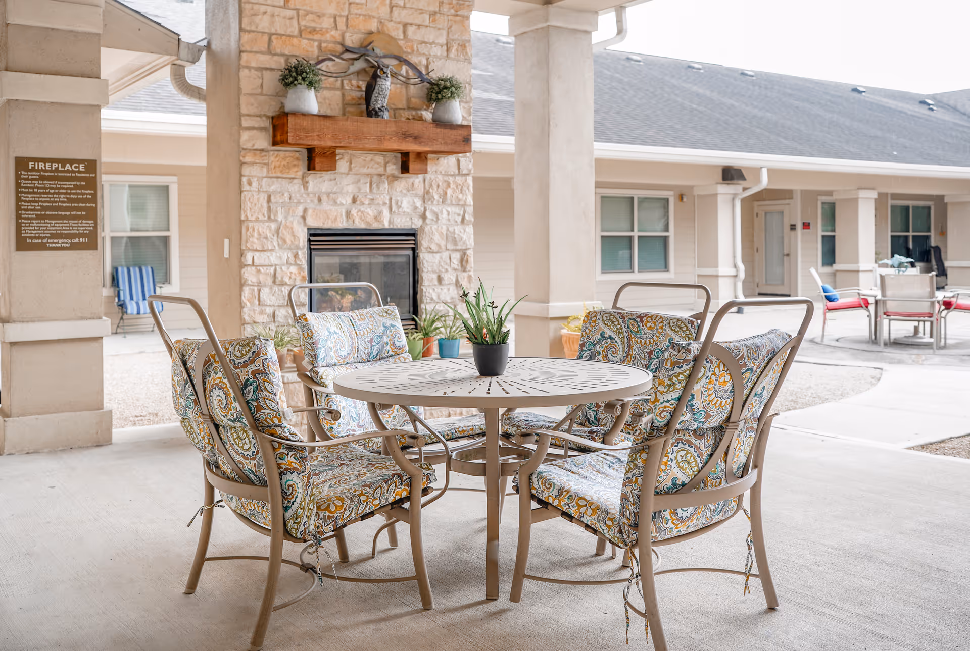 Outdoor covered seating area with a round metal table and four cushioned chairs featuring a colorful paisley pattern. Behind the table is a stone fireplace with a wooden mantel decorated with two small potted plants and a metal owl sculpture. The area is part of a senior living facility courtyard with additional seating and building entrances visible in the background.