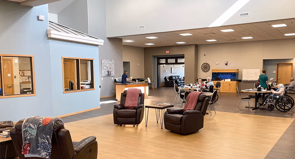 A spacious senior living facility common area with comfortable leather recliners, small tables, and several seniors seated at tables, some in wheelchairs. The room has high ceilings, light wood flooring, and a large fish tank mounted on the wall. A staff member is assisting residents, and there is a reception desk in the background.